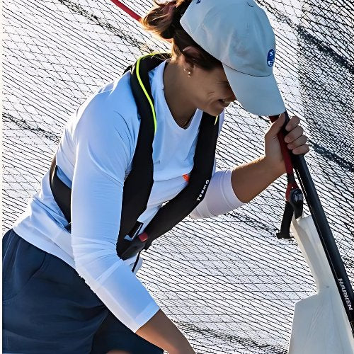 A person wearing a light blue cap, white long-sleeve shirt, blue shorts, and a life vest holds onto a pole with netting in the background, possibly engaged in a water-based activity like sailing.