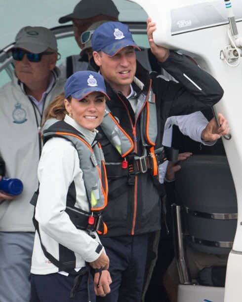 A man and a woman wearing blue caps and Cruising Solutions’ TeamO PFD with BackTow™ - Offshore Blue Hammar stand by a boat’s entrance, smiling, with several others in casual outdoor attire visible in the background.