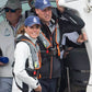 A man and a woman on a boat wear Cruising Solutions TeamO PFD with BackTow™ - Offshore Black ProSensor Elite, navy blue caps, and casual jackets. Both smile while looking outward; other casually dressed people wearing sunglasses are in the background.