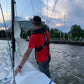 A person in a red shirt and a Cruising Solutions TeamO PFD with BackTow™ - Offshore Black ProSensor Elite stands on a sailboat handling the sail, with choppy water, buildings, trees, and cloudy skies in the background.