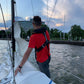 A person in a red shirt and a Cruising Solutions TeamO PFD with BackTow™ - Offshore Blue Hammar stands on a sailboat near a city shoreline, with buildings, trees, and a pedestrian bridge under a partly cloudy evening sky.
