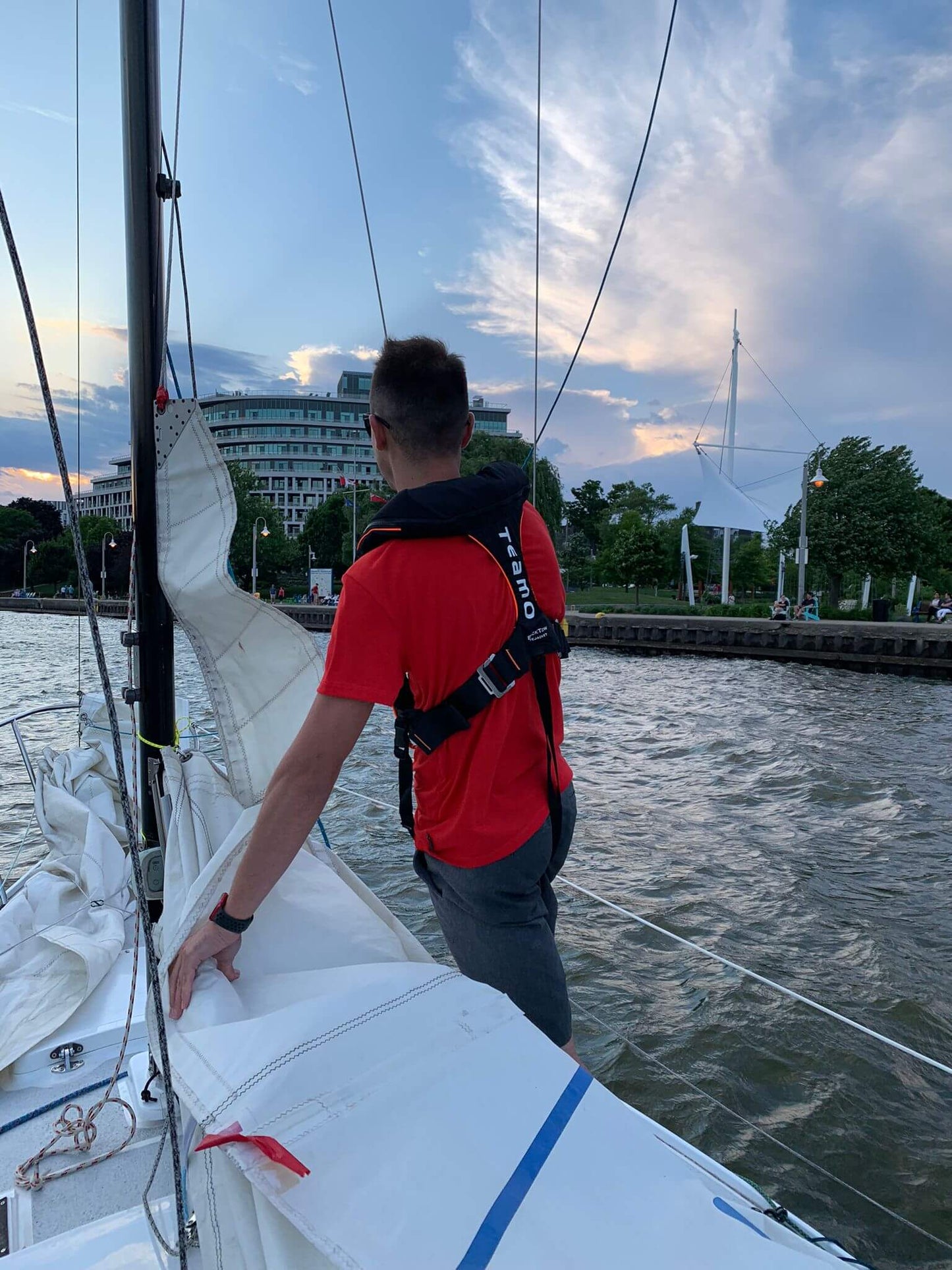 A person in a red shirt and a Cruising Solutions TeamO PFD with BackTow™ - Offshore Blue Hammar stands on a sailboat near a city shoreline, with buildings, trees, and a pedestrian bridge under a partly cloudy evening sky.