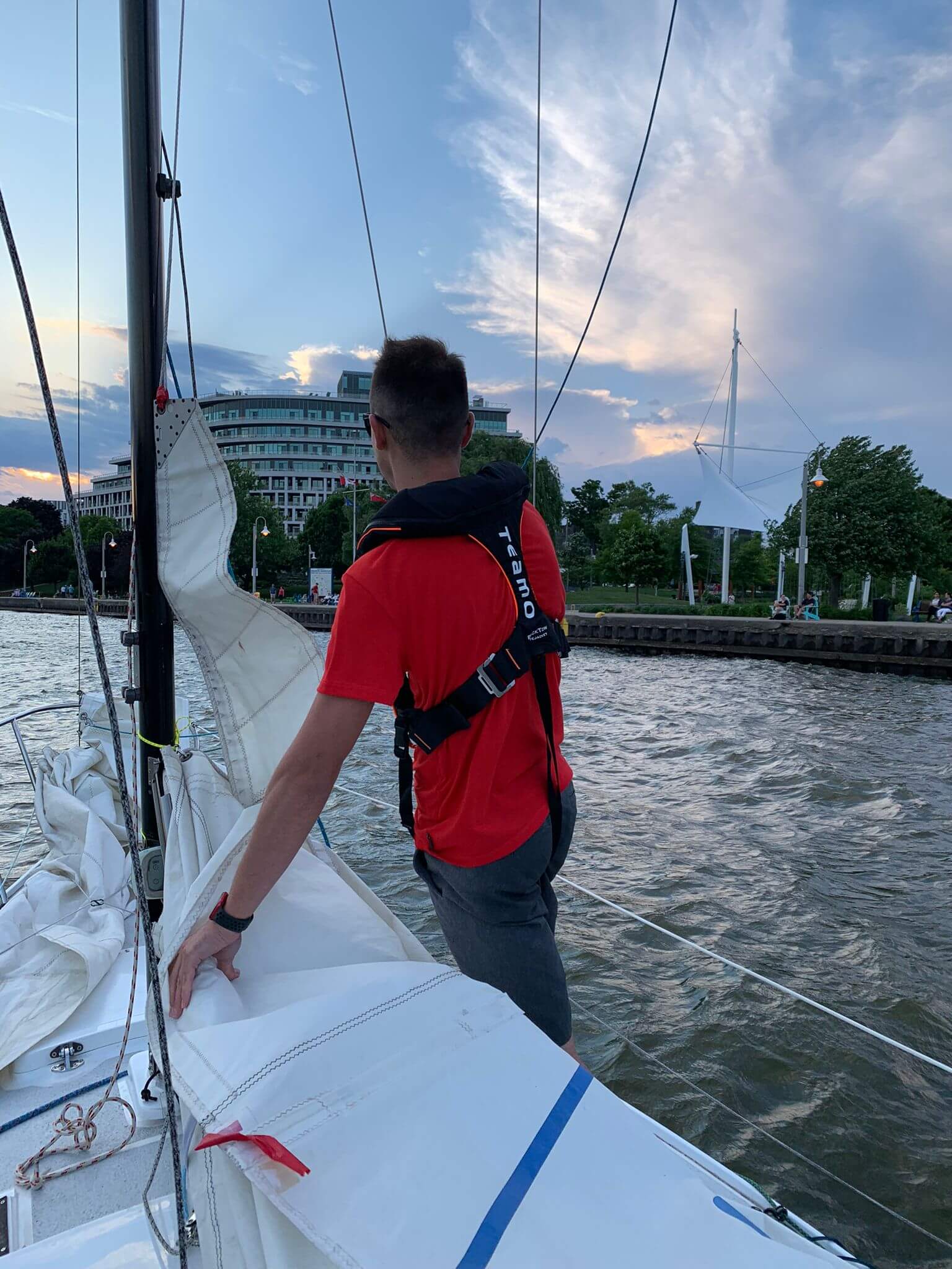 A person in a red shirt and a Cruising Solutions TeamO PFD with BackTow™ - Offshore Blue Hammar stands on a sailboat near a city shoreline, with buildings, trees, and a pedestrian bridge under a partly cloudy evening sky.