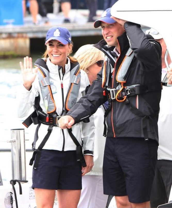 Two people on a dock wear Cruising Solutions' TeamO PFD with BackTow™ - Ocean Grey ProSensor Elite, navy shorts, and caps. One smiles and waves while the other adjusts their cap, with others and a boat seen in the background.