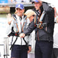Two people in Cruising Solutions TeamO PFDs with BackTow™ (Ocean Black ProSensor Elite), hats, and shorts stand on a dock by a boat—one waves and smiles, the other adjusts their cap. Others and water are visible in the background.