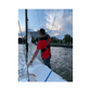 A person in a red shirt and the Cruising Solutions TeamO PFD with BackTow™ - Offshore Black Hammar stands on a sailboat holding the sail, with buildings, trees, and a cloudy sky in the background.