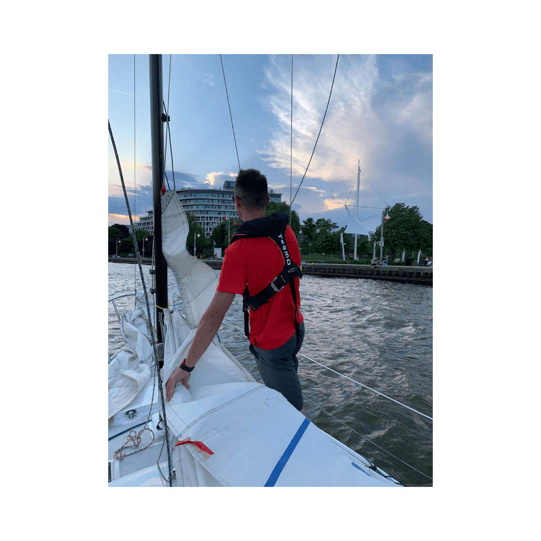 A person in a red shirt and the Cruising Solutions TeamO PFD with BackTow™ - Offshore Black Hammar stands on a sailboat holding the sail, with buildings, trees, and a cloudy sky in the background.