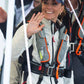 A woman in a light jacket and Cruising Solutions' TeamO PFD with BackTow™ - Offshore Grey Hammar waves and smiles. She also wears a navy cap with an emblem, joined by others in similar offshore inflatable PFDs.