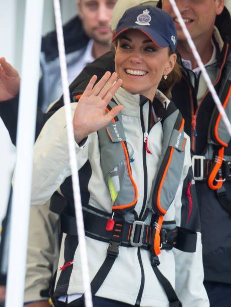 A woman in a light jacket and Cruising Solutions' TeamO PFD with BackTow™ - Offshore Grey Hammar waves and smiles. She also wears a navy cap with an emblem, joined by others in similar offshore inflatable PFDs.