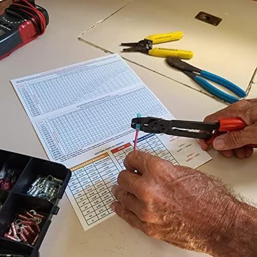 Hands expertly maneuver a wire stripper on a red wire above a table, with a printed chart lying beside a toolbox and pliers. The Boat Galleys 12 Volt Wiring Size Quick Reference serves as an indispensable reference for any 12 volt wiring project.